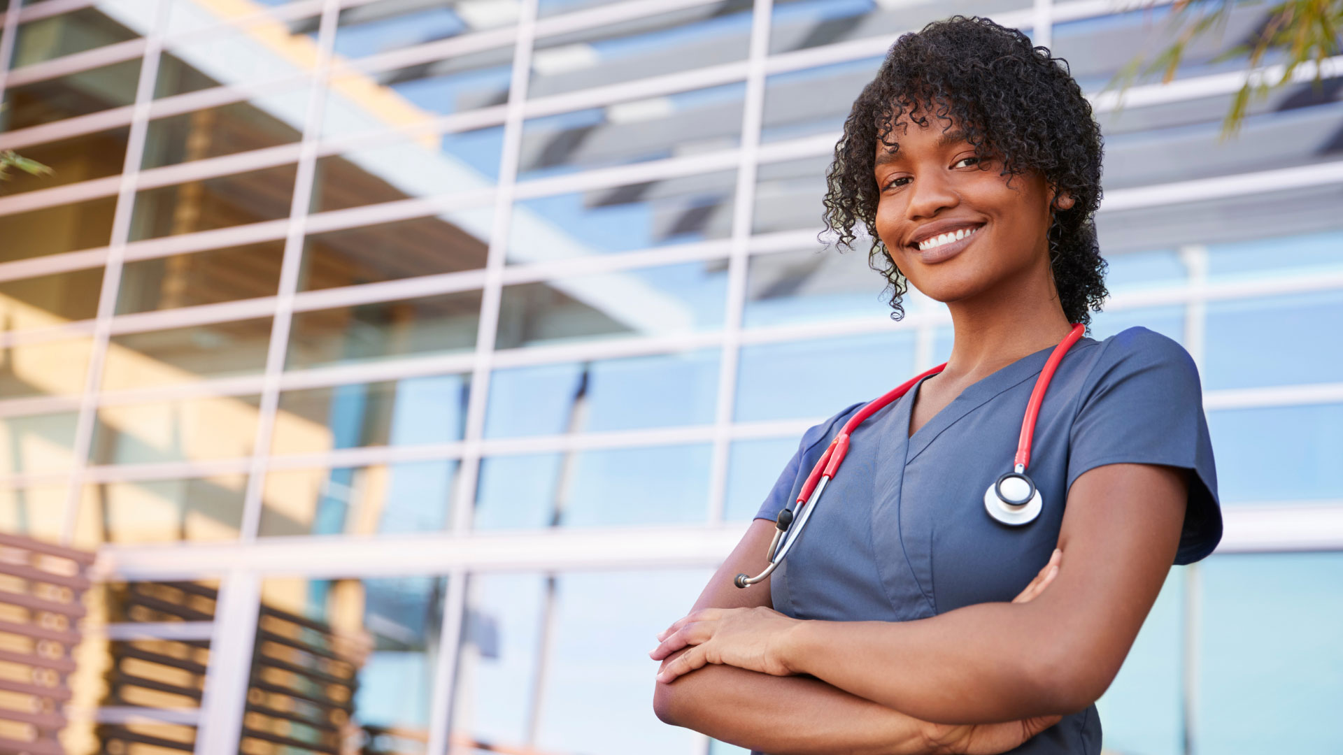 Healthcare professional wearing blue scrubs and a red stethoscope standing confidently with arms crossed in front of an ambulatory surgery center